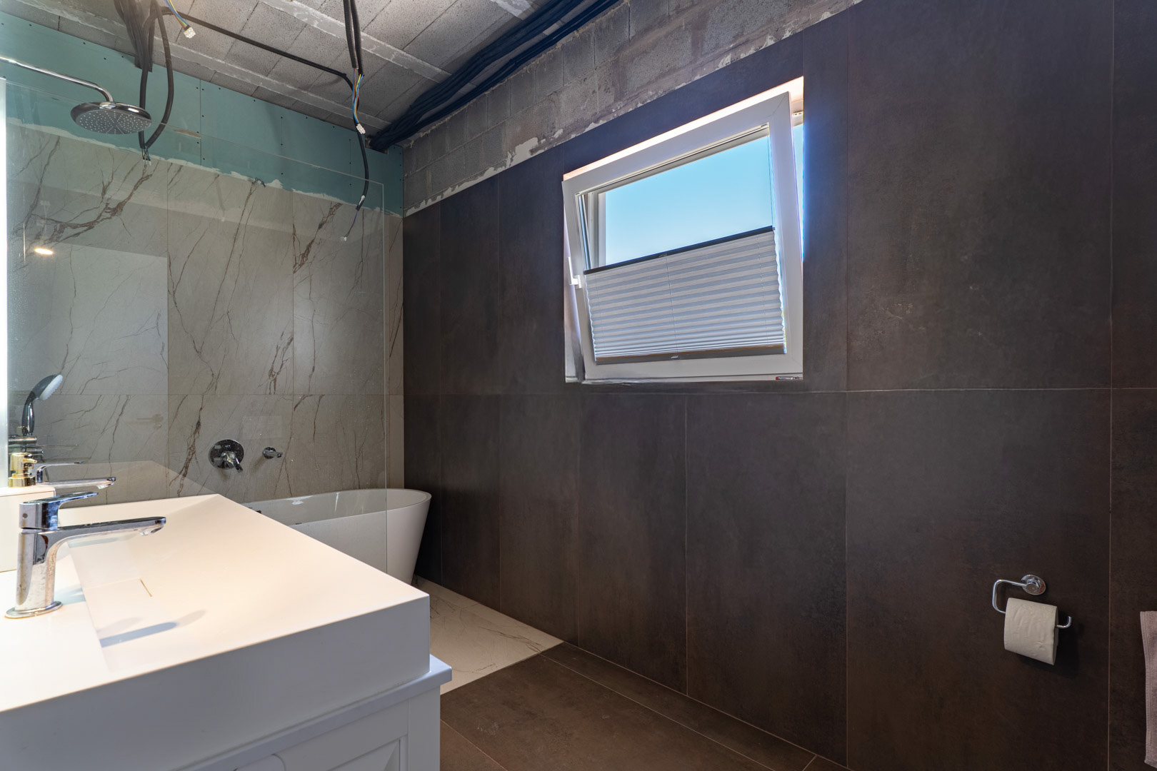 Salle de bain en cours de finition avec double vasque, baignoire îlot et douche à l'italienne. Les grands carreaux sombres contrastent avec le marbre et la fenêtre oscillo-battante.