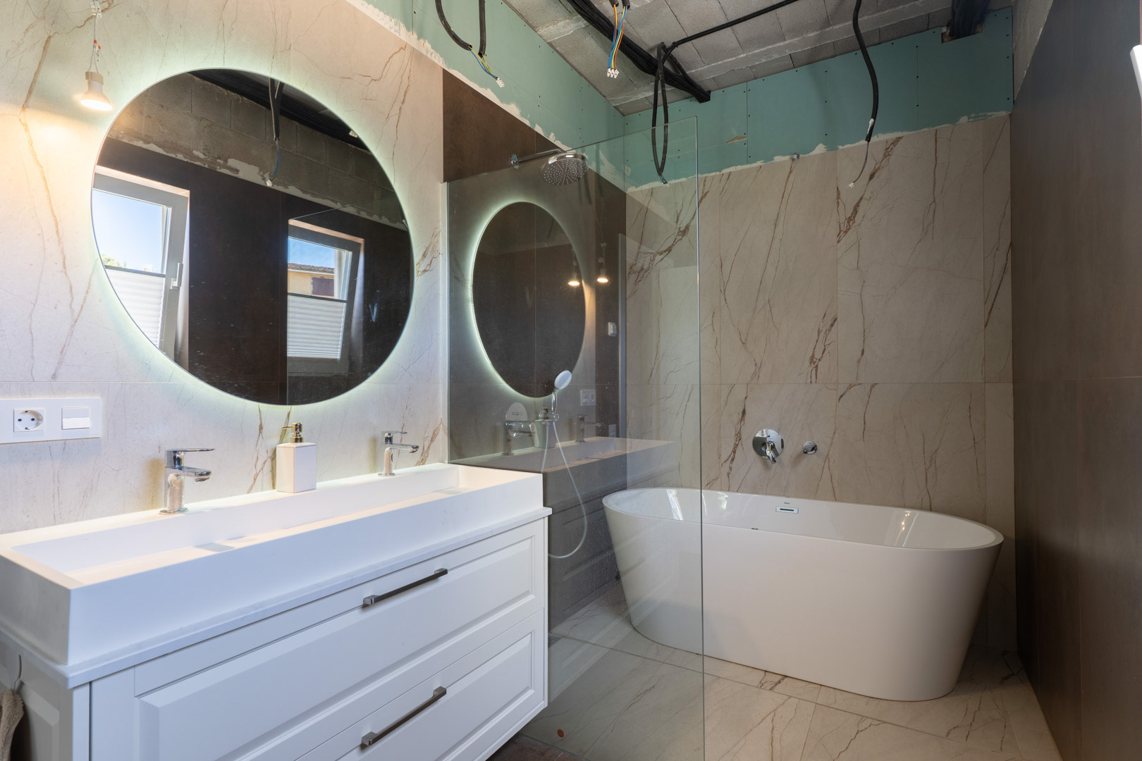 Salle de bain en cours de finition avec double vasque, miroir circulaire rétroéclairé et baignoire îlot sur grès cérame effet marbre.