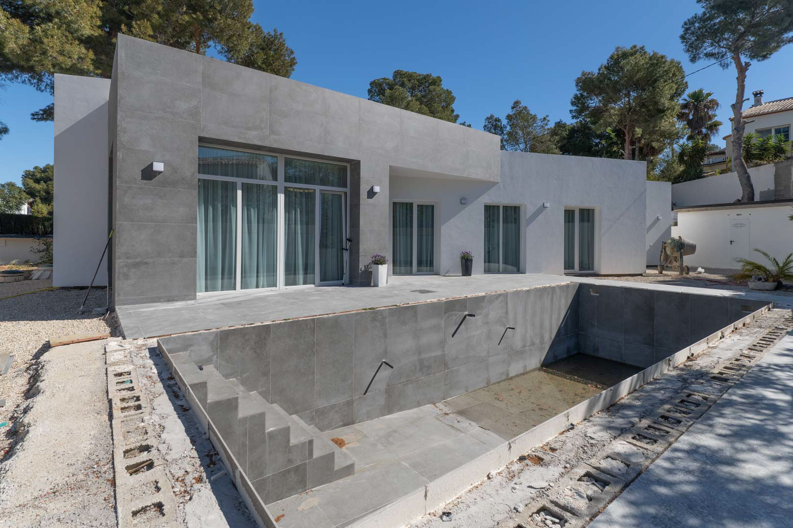 Villa minimaliste de plain-pied avec façade en grès cérame gris et piscine en cours de finition, entourée de pins méditerranéens.