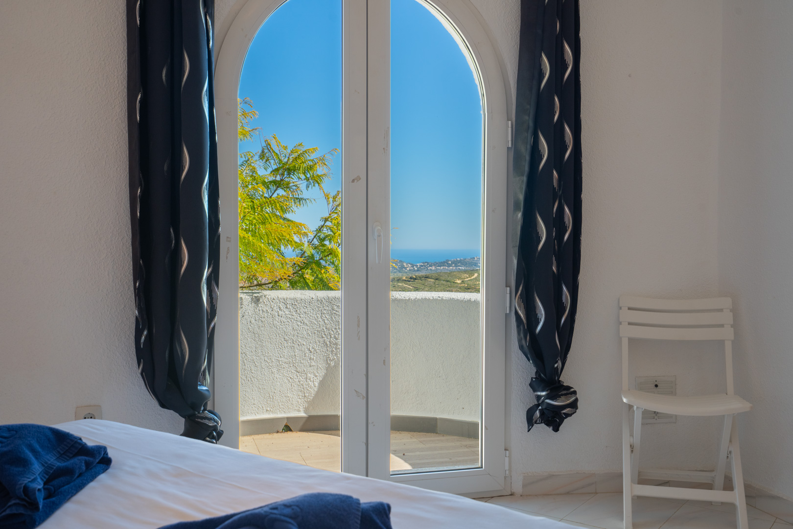 Chambre avec porte vitrée en plein cintre s'ouvrant sur un balcon privé avec vue sur la mer. Comprend des murs blancs et des rideaux sombres à motifs.