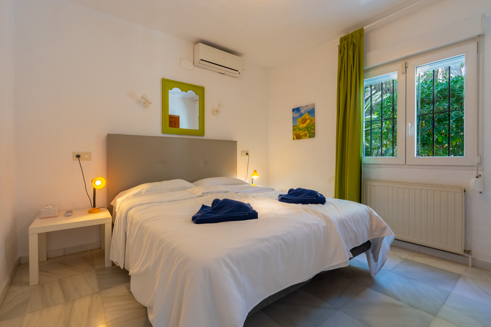 Chambre avec sol en marbre, climatisation et chauffage. Elle comprend une fenêtre avec grilles de sécurité, des rideaux verts et un miroir décoratif assorti.