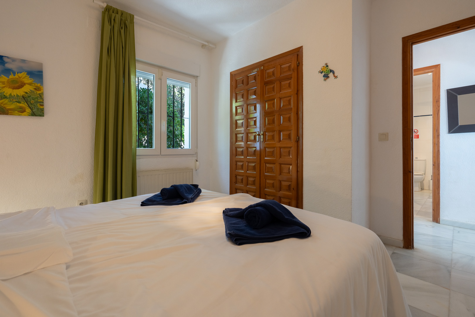 Chambre lumineuse avec armoire encastrée en bois sculpté, rideaux verts et accès à une salle de bain attenante avec sol en marbre.