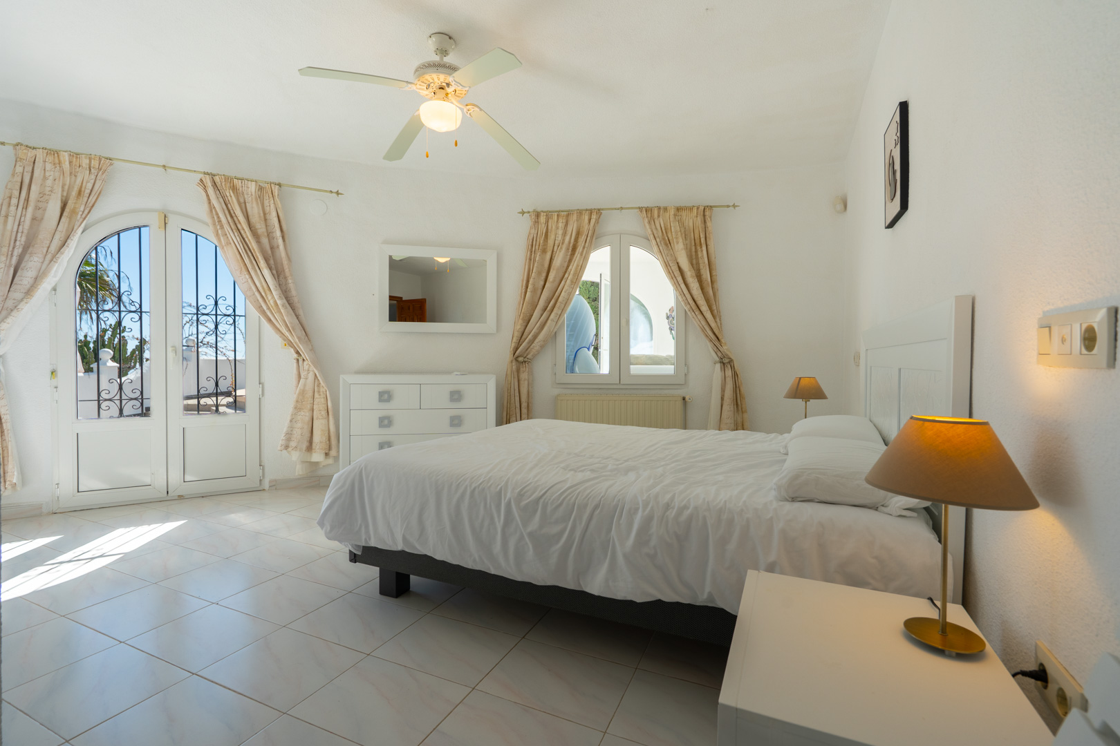 Chambre lumineuse avec sol en grès, ventilateur de plafond et accès direct à l'extérieur par une porte vitrée cintrée avec ferronnerie décorative.