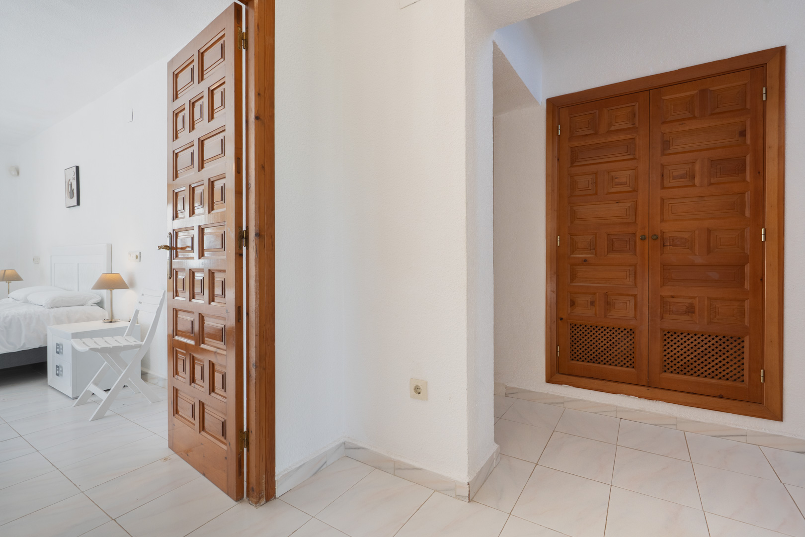 Couloir avec des portes en bois sculpté de style traditionnel et des placards intégrés, menant à une chambre lumineuse avec un sol en carrelage blanc.