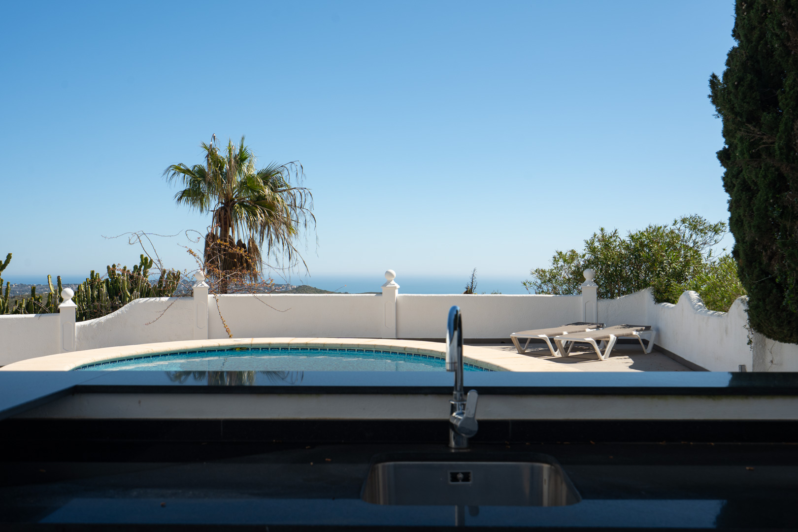 Cuisine d'été avec plan de travail noir et évier, offrant une vue sur la piscine arrondie, la terrasse ensoleillée et l'horizon marin.