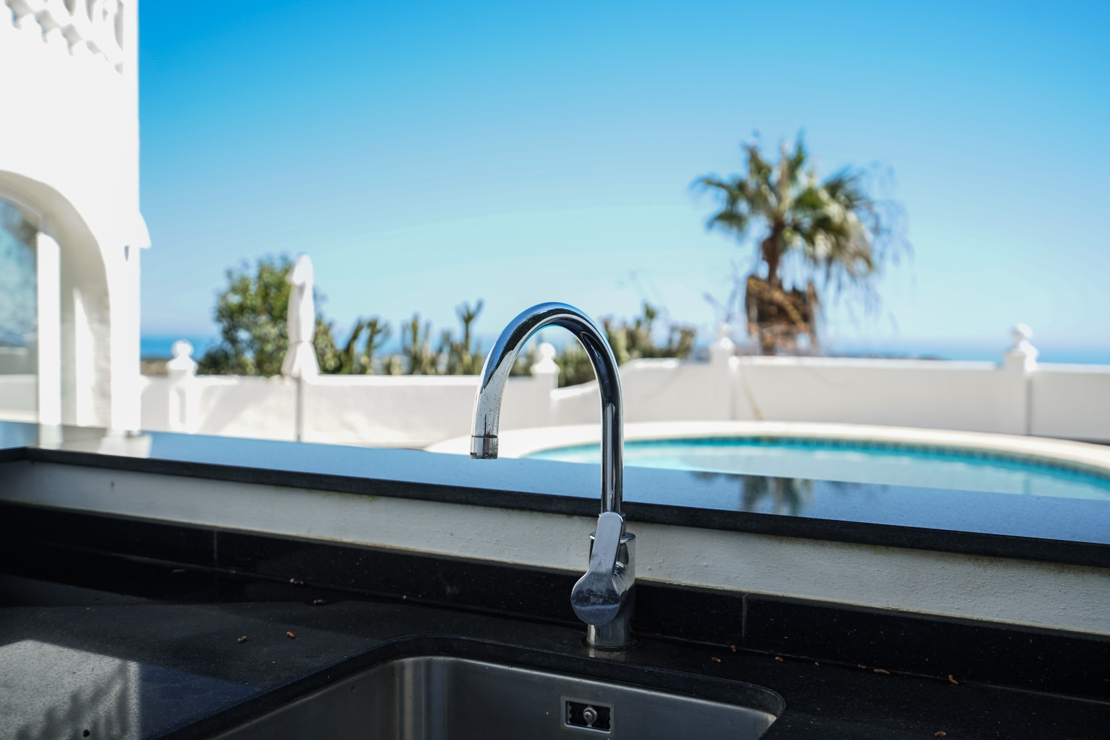 Cuisine extérieure équipée d'un plan de travail en granit noir et d'un évier en inox, avec vue sur la piscine et les jardins.