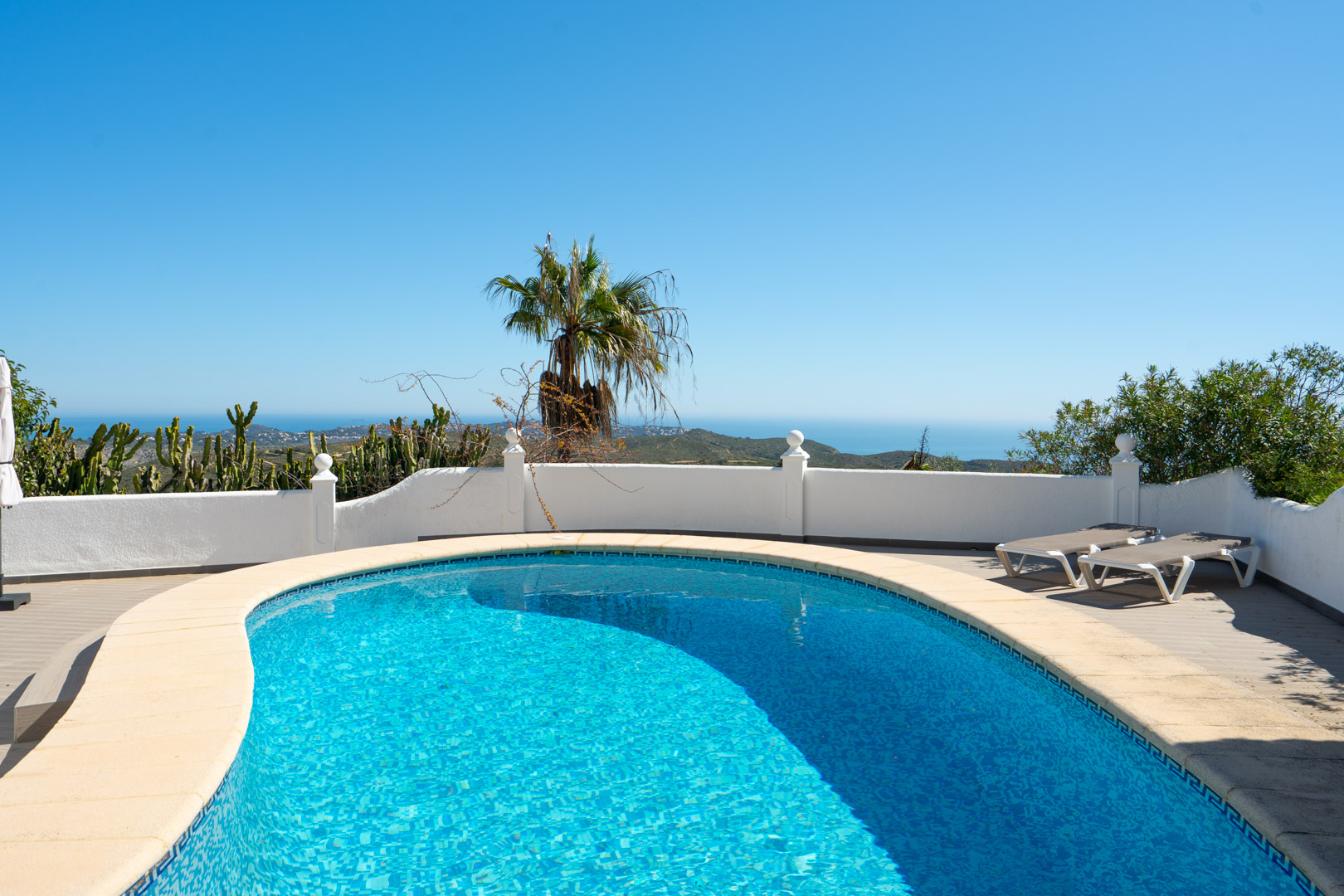 Piscine aux formes arrondies offrant une vue panoramique sur la mer, entourée d'une terrasse ensoleillée avec chaises longues et murets blancs.