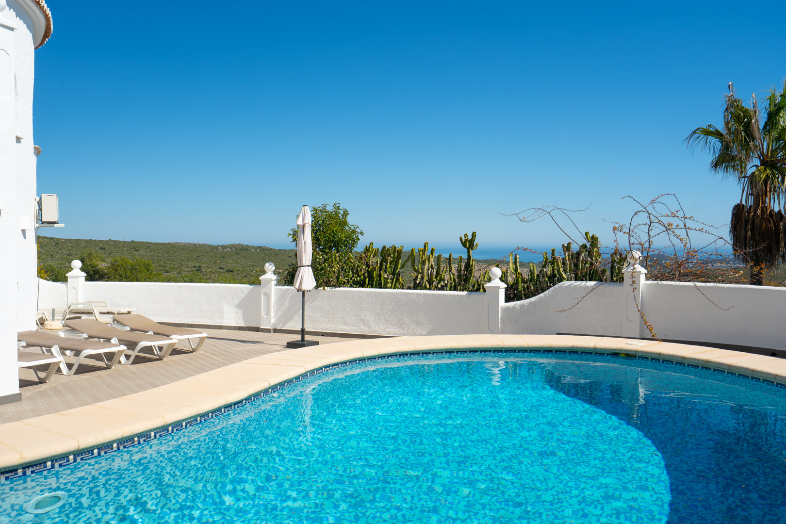 Piscine de forme courbe entourée d'une terrasse ensoleillée avec transats, offrant une vue panoramique sur la mer Méditerranée et la nature environnante.