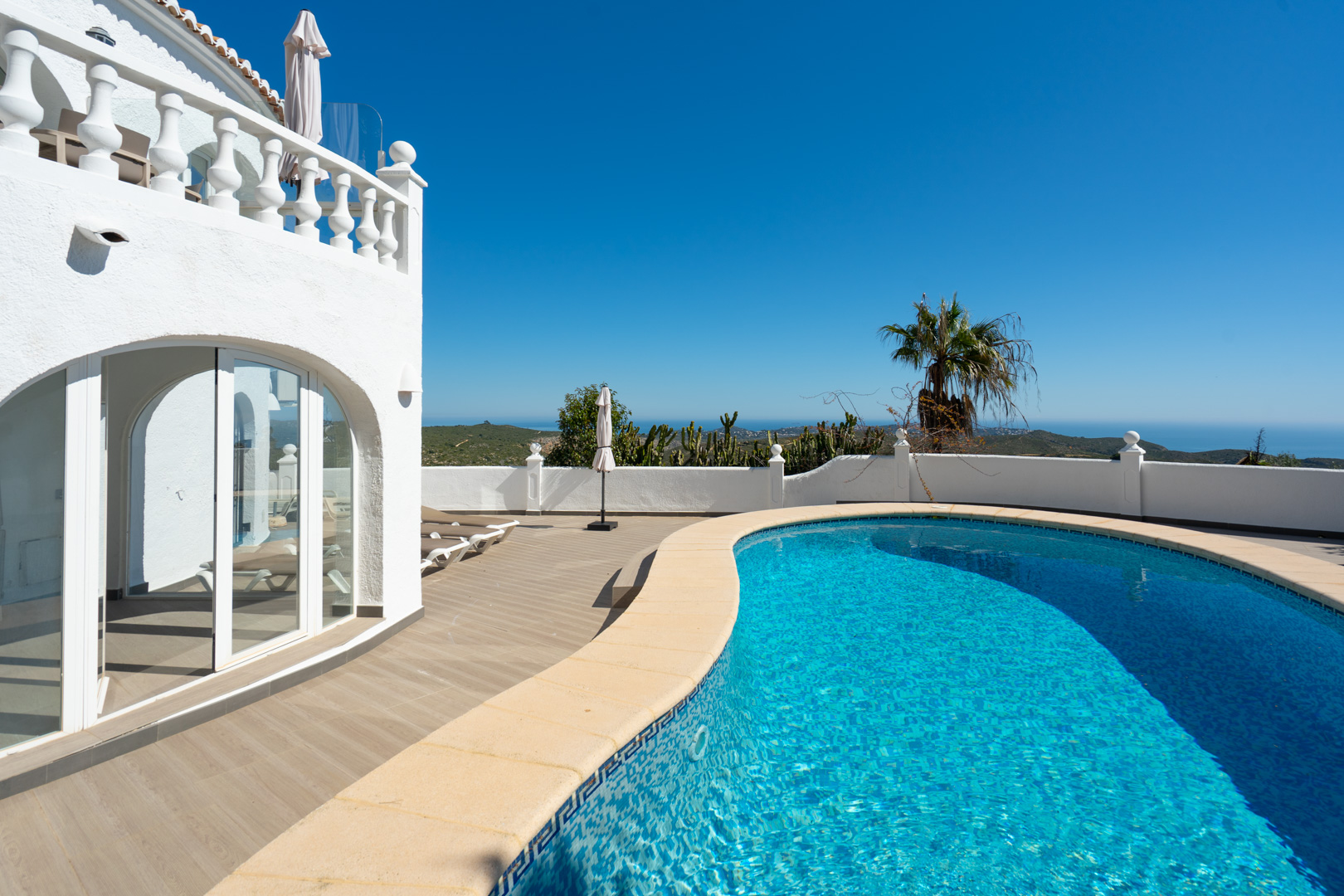 Piscine de forme libre avec terrasse en grès cérame imitation bois, offrant une vue dégagée sur la mer et un accès direct à la naya vitrée de la villa.