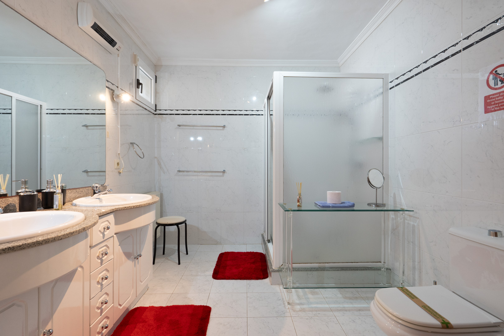 Salle de bain avec double vasque sur plan en granit, grand miroir et douche avec paroi en verre dépoli. Comprend une climatisation split et des murs carrelés.