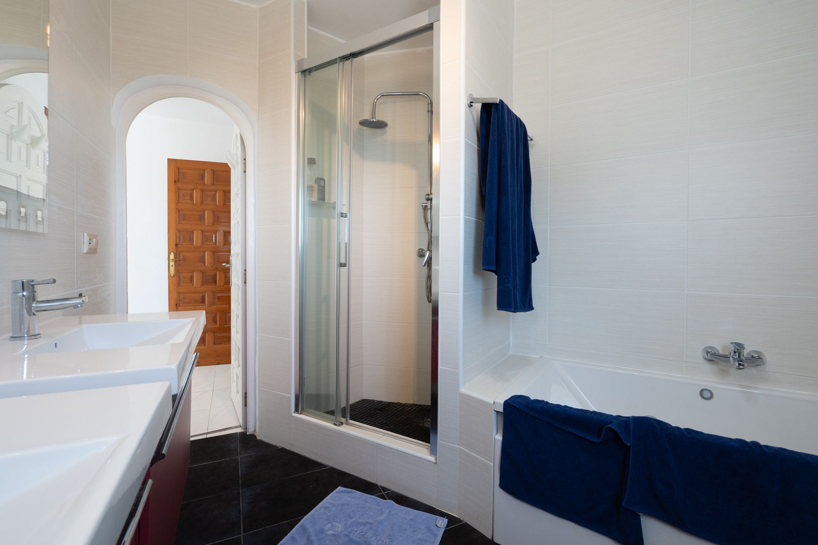 Salle de bain comprenant un double lavabo, une baignoire et une douche séparée avec paroi vitrée. Carrelage mural clair et sol sombre contrasté avec porte voûtée.