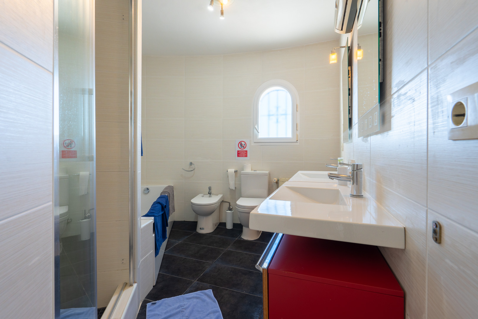 Salle de bain équipée d'une double vasque sur meuble rouge, d'un bidet, de toilettes et d'une baignoire. Murs carrelés clairs et petite fenêtre cintrée.