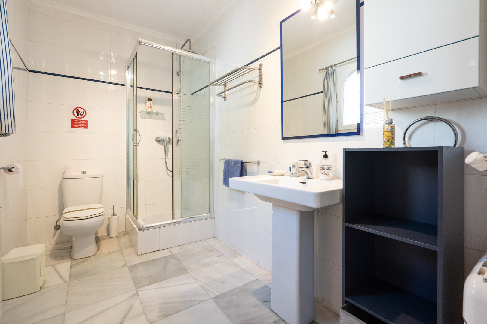 Salle de bains avec carrelage mural blanc, sol en marbre, cabine de douche en verre et lavabo sur colonne avec miroir au cadre bleu.