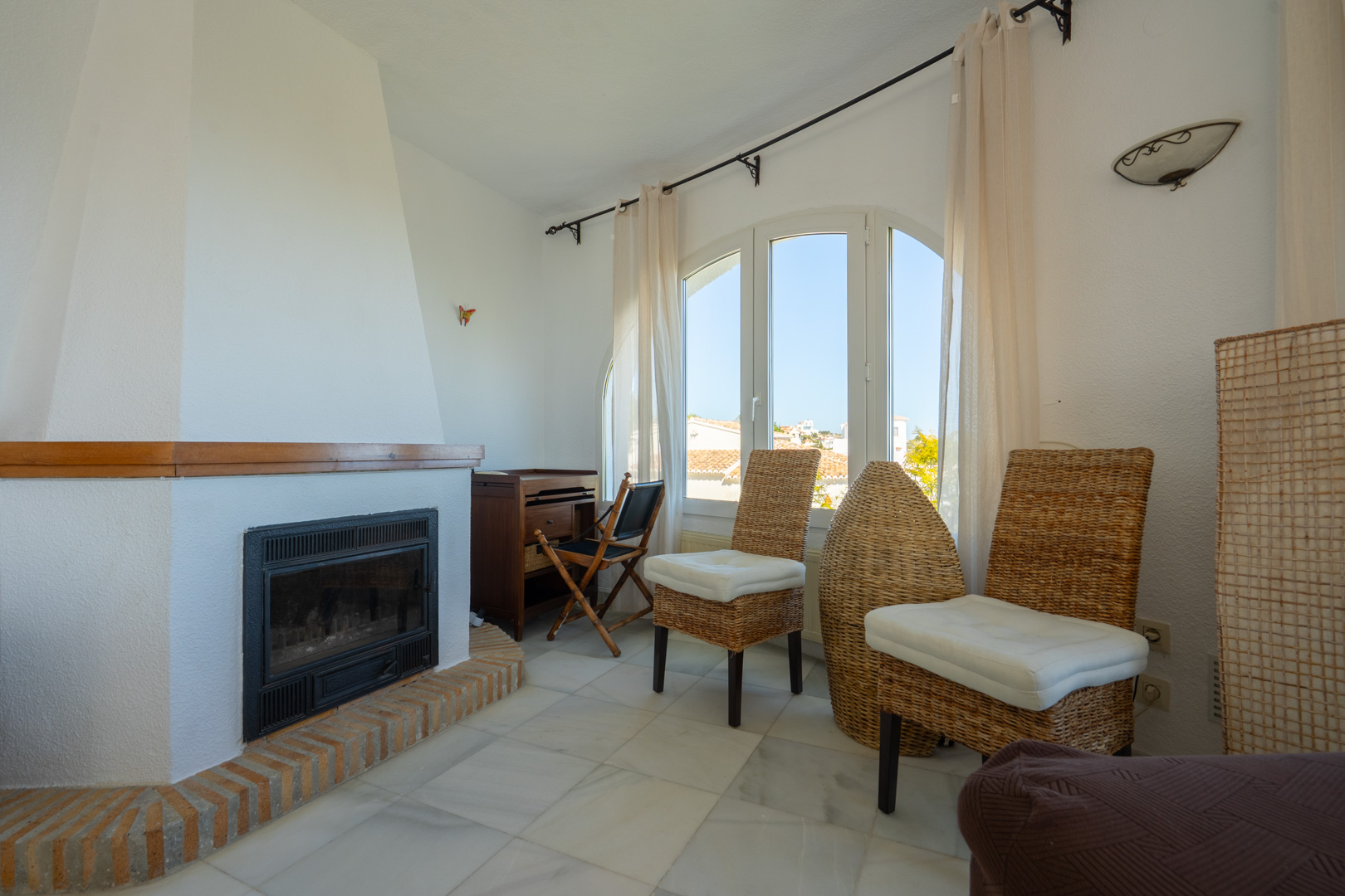 Salon avec cheminée encastrée sur socle en briques, sol en marbre et fenêtre cintrée offrant une lumière naturelle sur le mobilier en osier traditionnel.