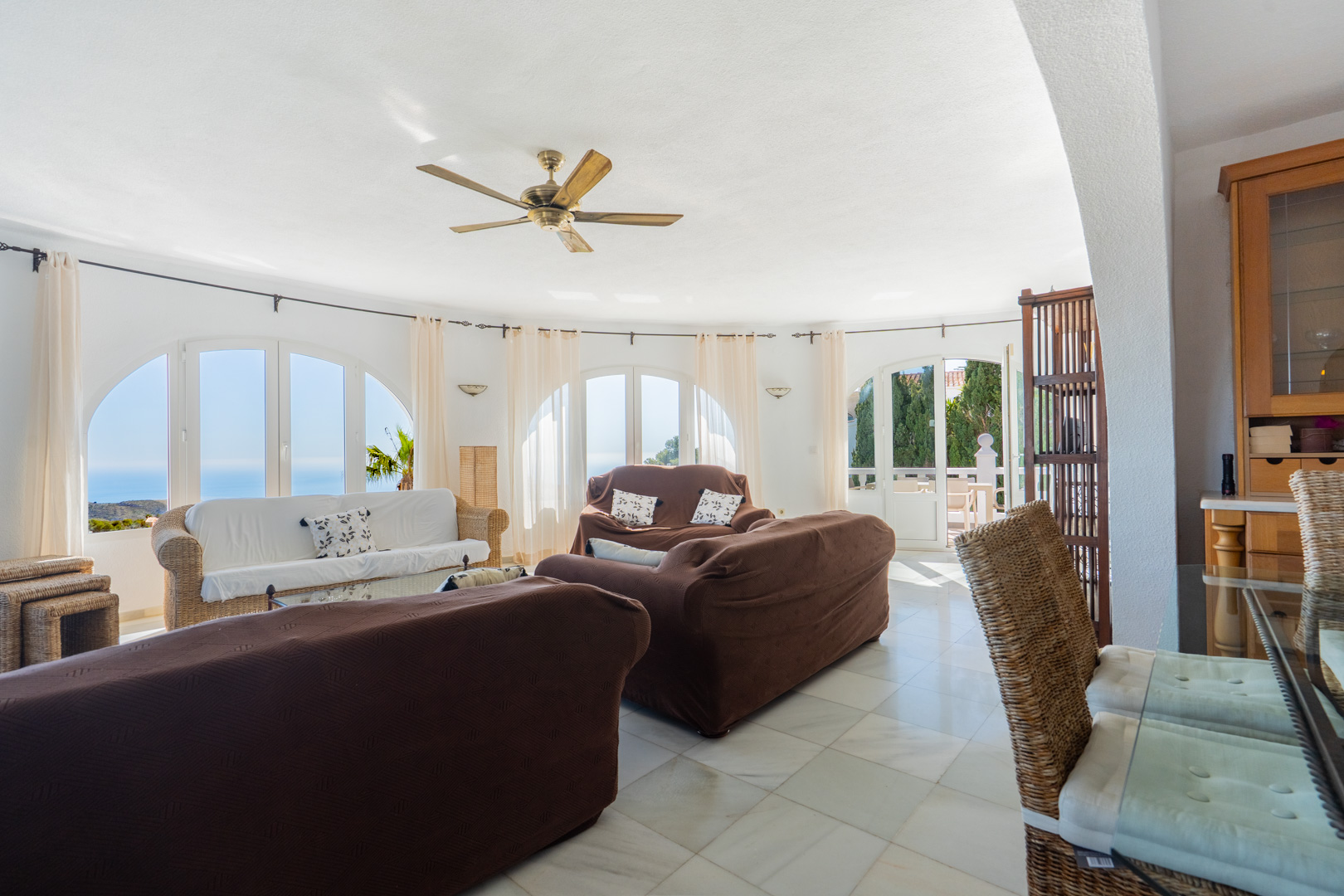Salon circulaire avec fenêtres en arc offrant une vue sur la mer, sol en marbre clair, ventilateur de plafond et mobilier en osier traditionnel.