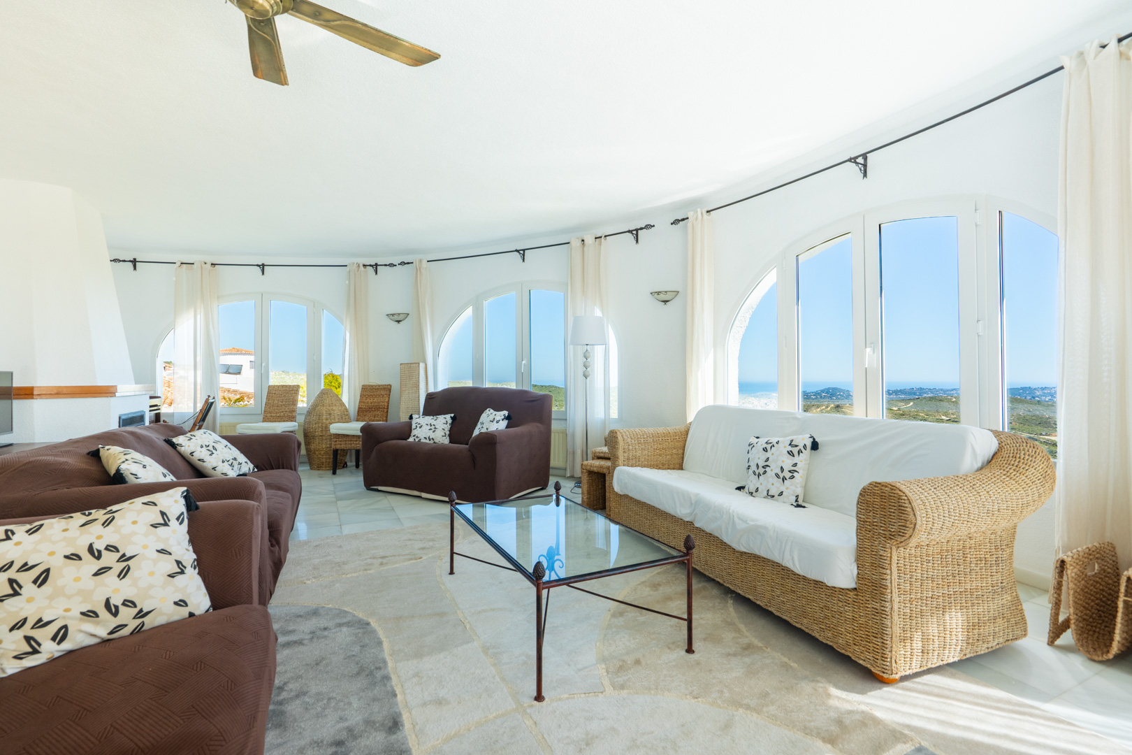 Salon circulaire avec fenêtres en arche offrant une vue sur mer, mobilier en osier et tissu, et une cheminée d'angle sous un ventilateur.