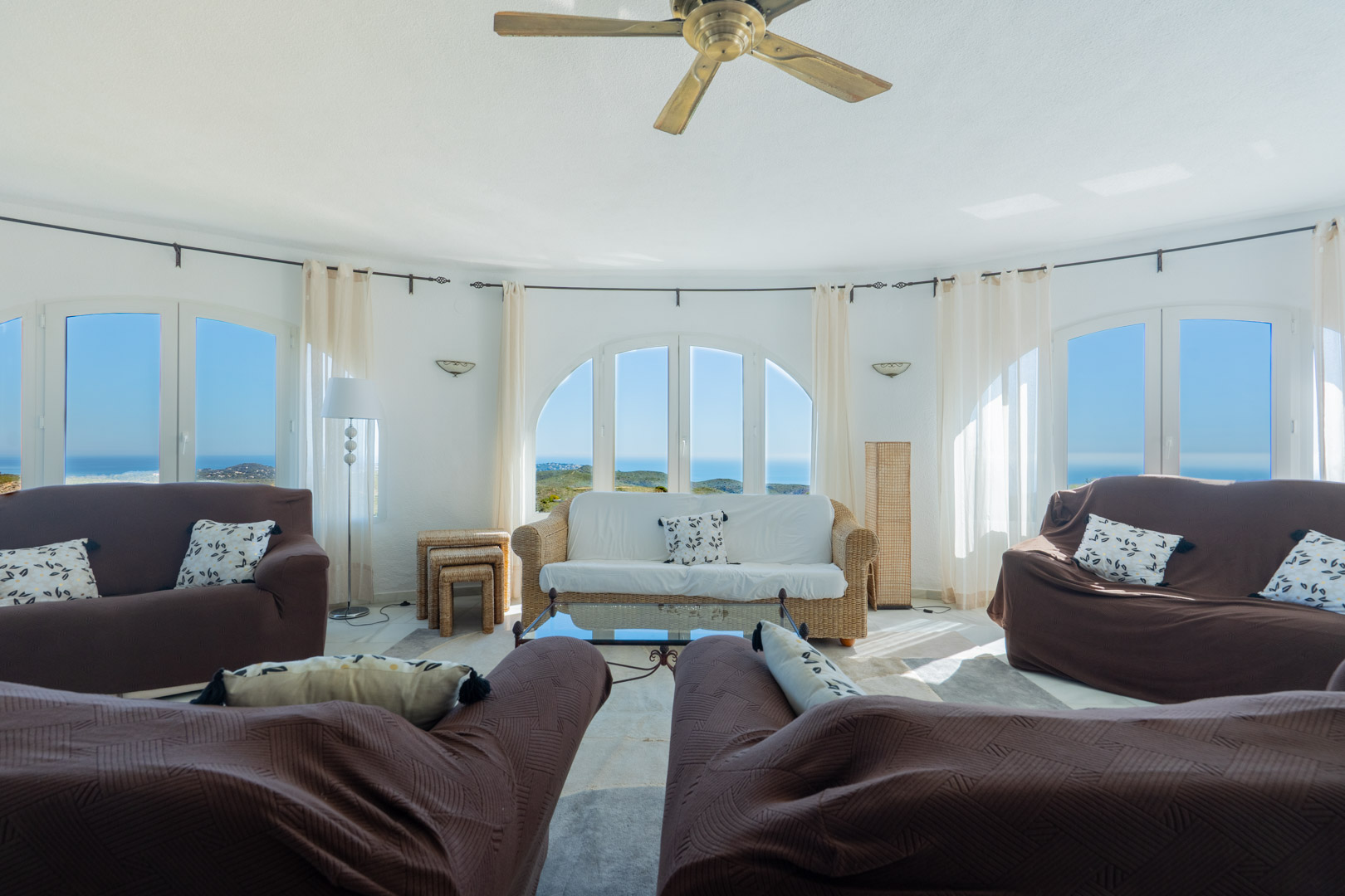 Salon circulaire doté de fenêtres en arc offrant une vue panoramique sur la mer. Comprend un ventilateur de plafond, du mobilier en osier et une lumière naturelle.