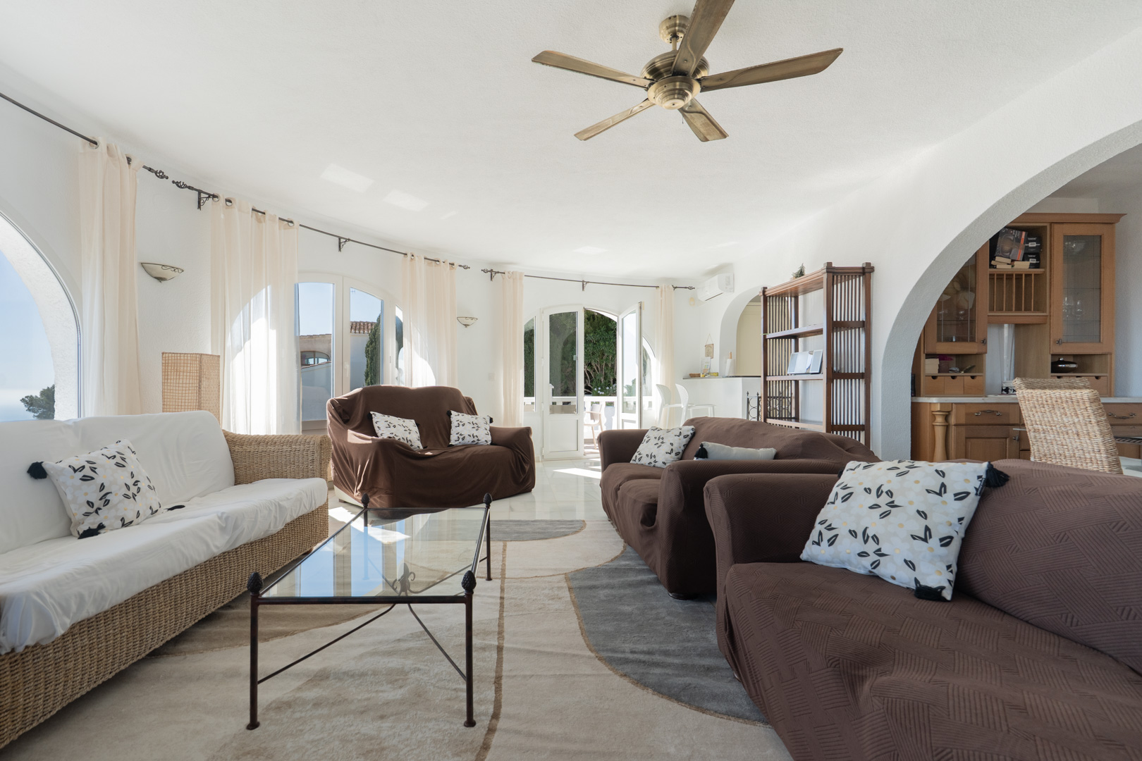 Salon lumineux avec mur incurvé, fenêtres en arc et accès terrasse. Comprend un ventilateur de plafond en bois, un sol carrelé et une ouverture voûtée vers la cuisine.