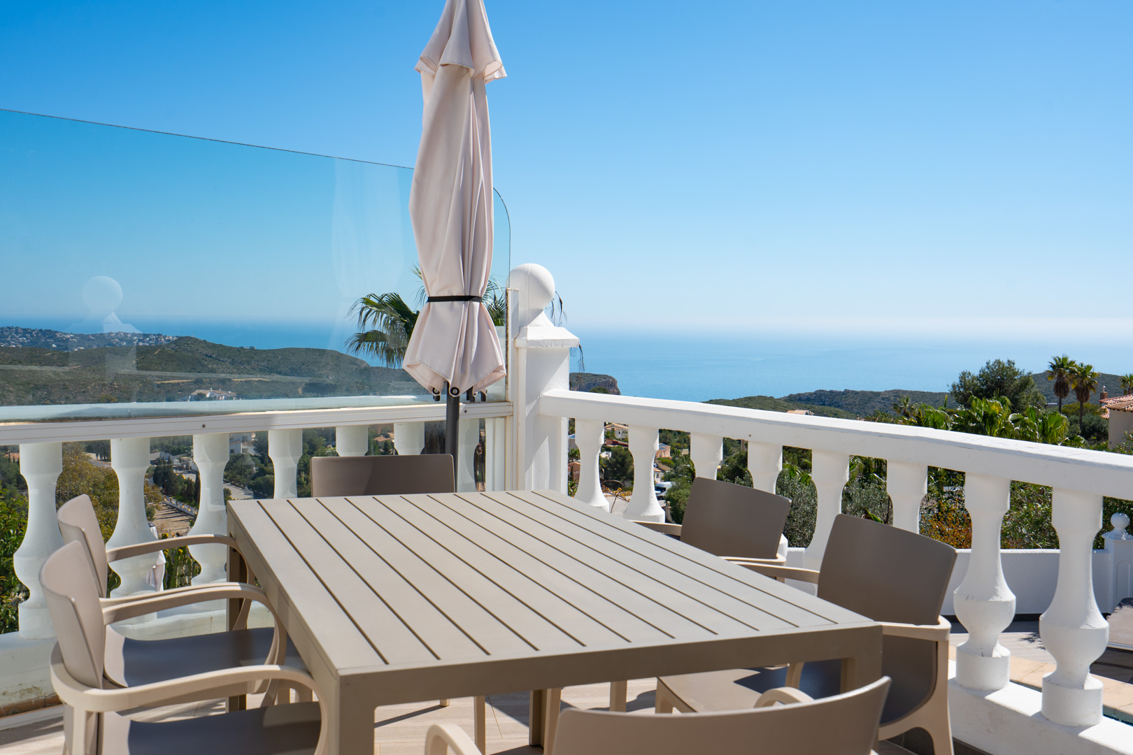 Terrasse de repas extérieure avec table et chaises, bordée d'une balustrade blanche et de parois vitrées offrant une vue panoramique sur la mer Méditerranée.