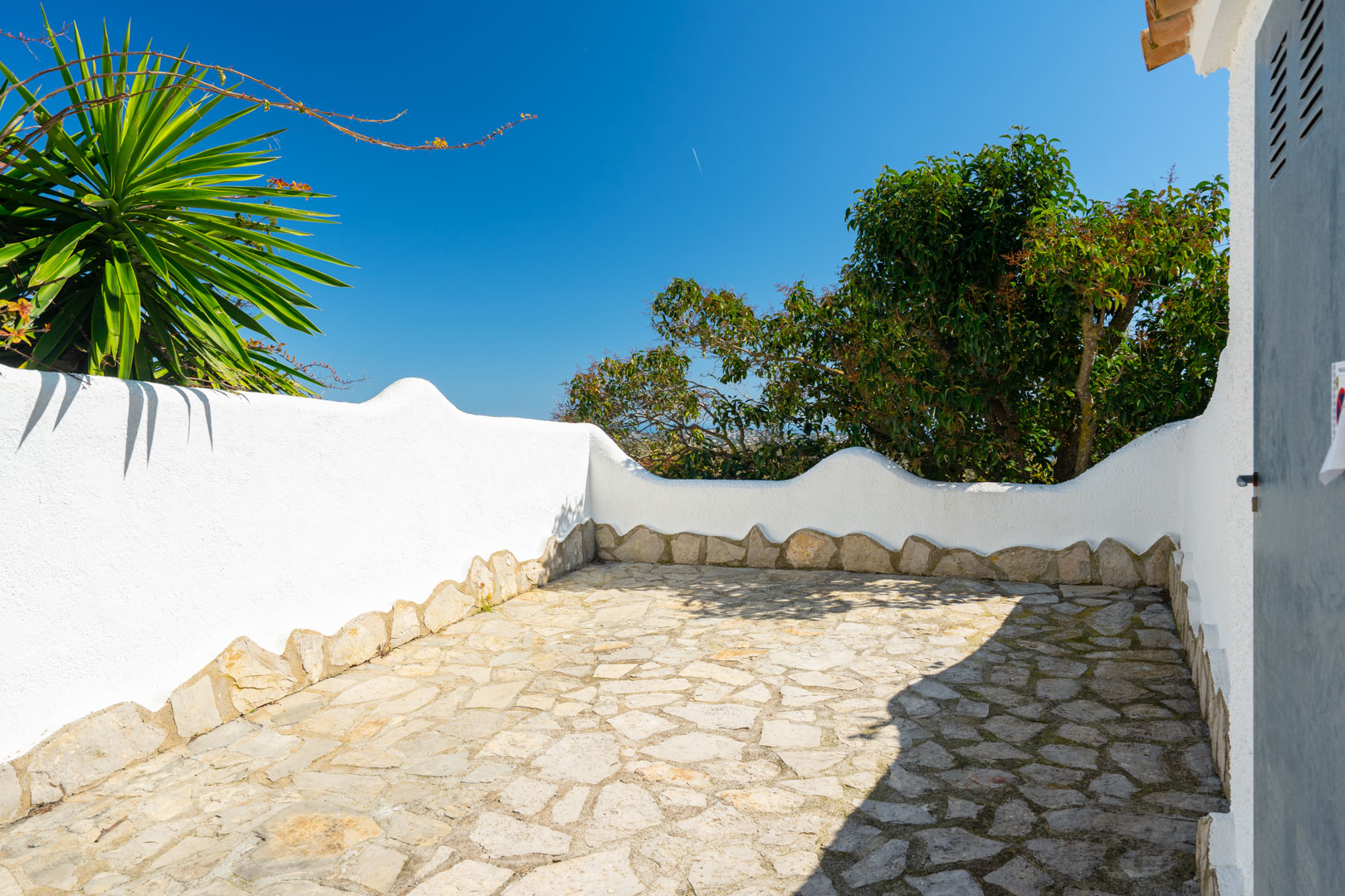Terrasse extérieure avec sol en pierre naturelle irrégulière et murs blancs aux formes ondulées, entourée de plantes méditerranéennes.