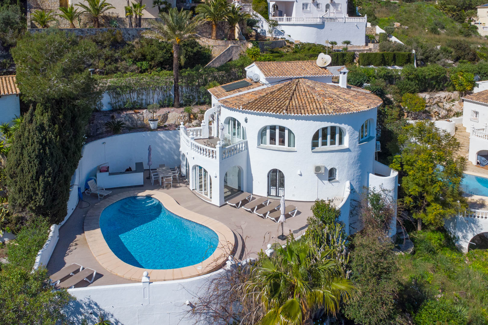 Vue aérienne de la villa avec piscine en forme de rein, grande terrasse ensoleillée, façade blanche traditionnelle et panneaux solaires sur le toit en tuiles.