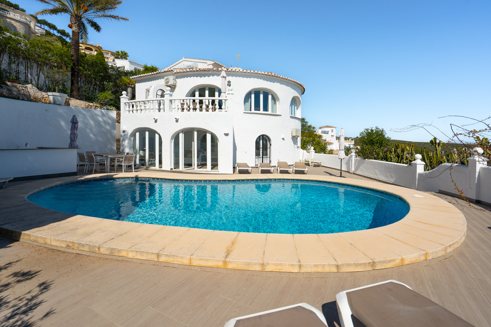Vue extérieure de la villa avec piscine de forme libre, grande terrasse en grès cérame effet bois et façade blanche aux fenêtres cintrées et balustrade classique.
