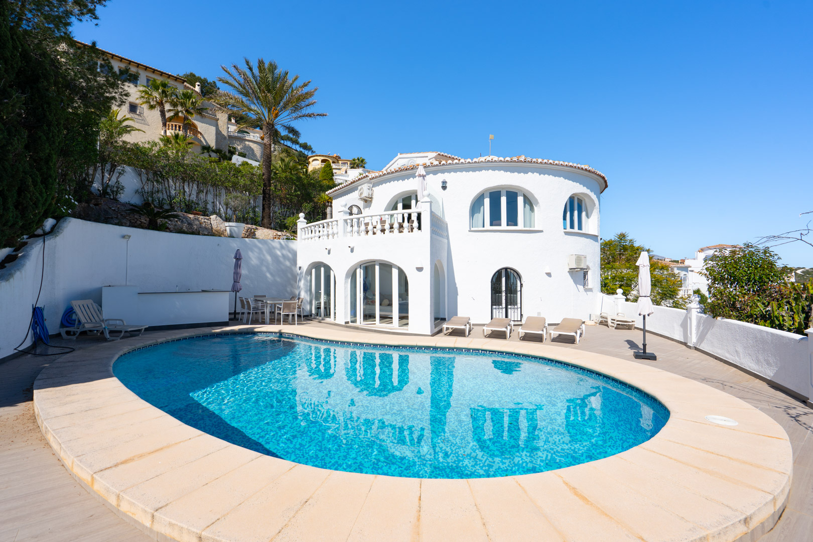 Vue extérieure de la villa avec piscine ovale, vaste terrasse ensoleillée et façade blanche traditionnelle dotée d'arches et d'une naya vitrée.