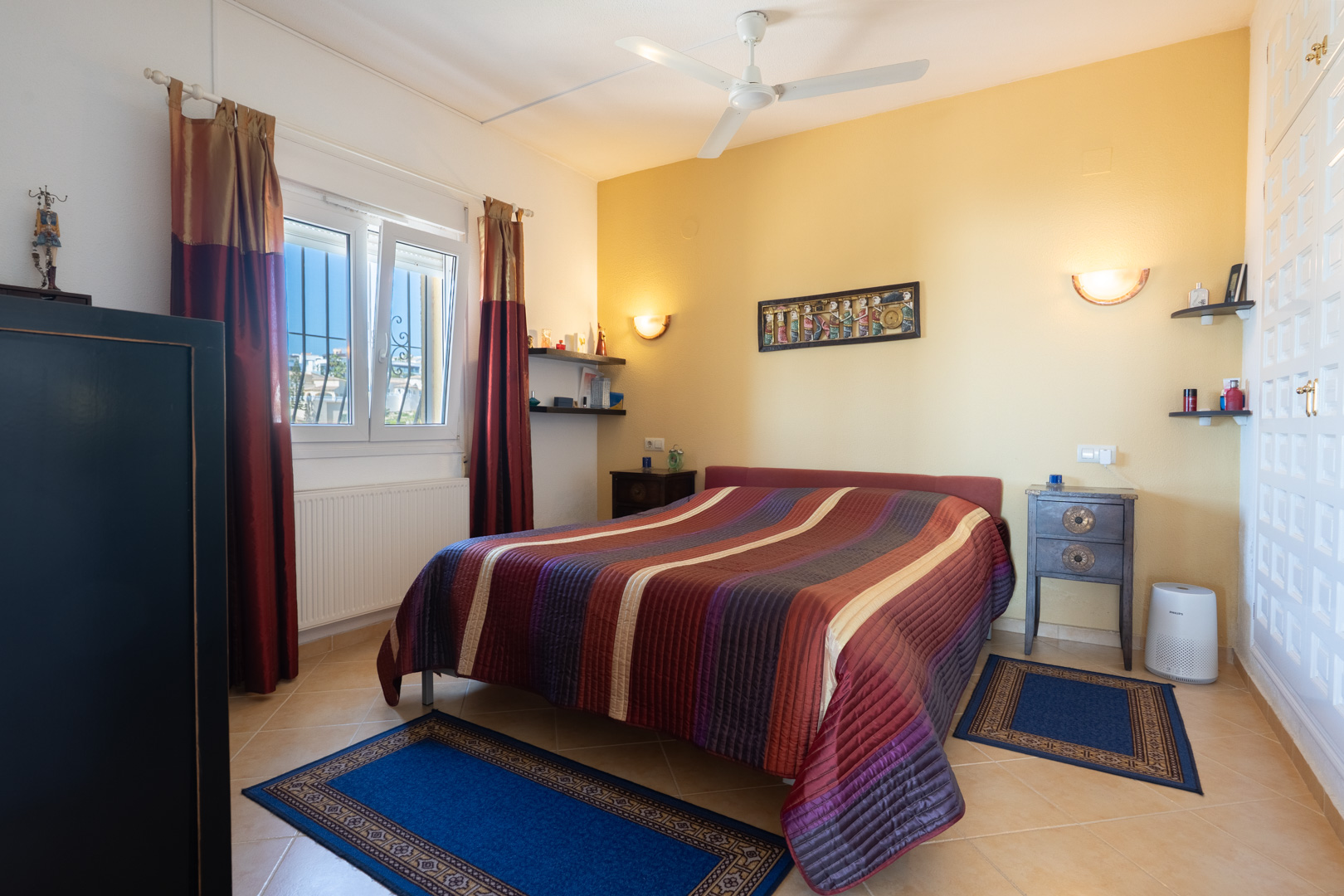 Chambre avec lit double, mur d'accent jaune, ventilateur de plafond et placards intégrés blancs. Équipée d'un chauffage par radiateur et de carrelage.