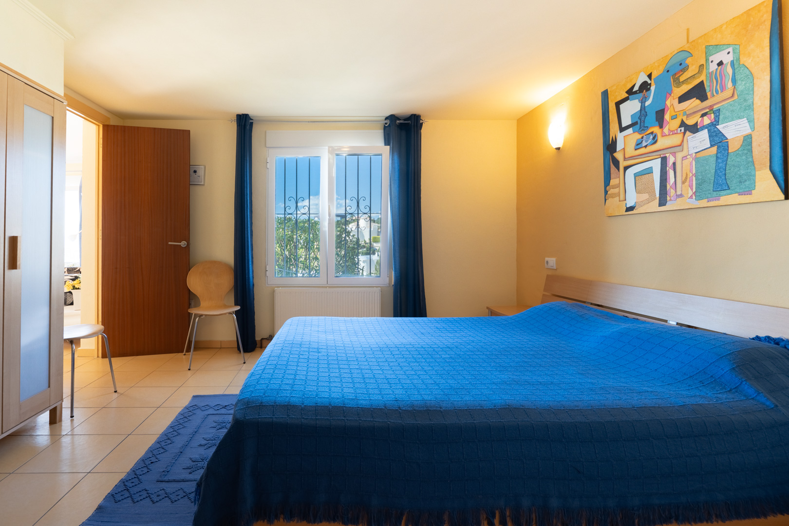 Chambre avec lit double, textiles bleus, armoire en bois et carrelage clair. Une fenêtre avec grilles de sécurité apporte une luminosité naturelle.