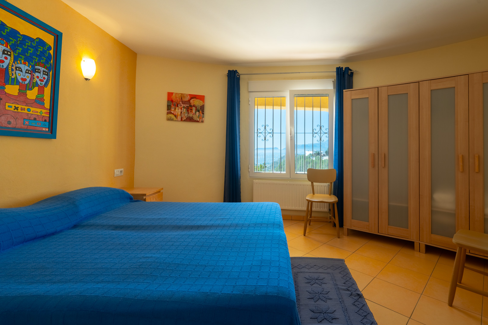Chambre lumineuse avec lit double, murs ocre, armoire en bois à panneaux translucides et fenêtre offrant une vue panoramique.