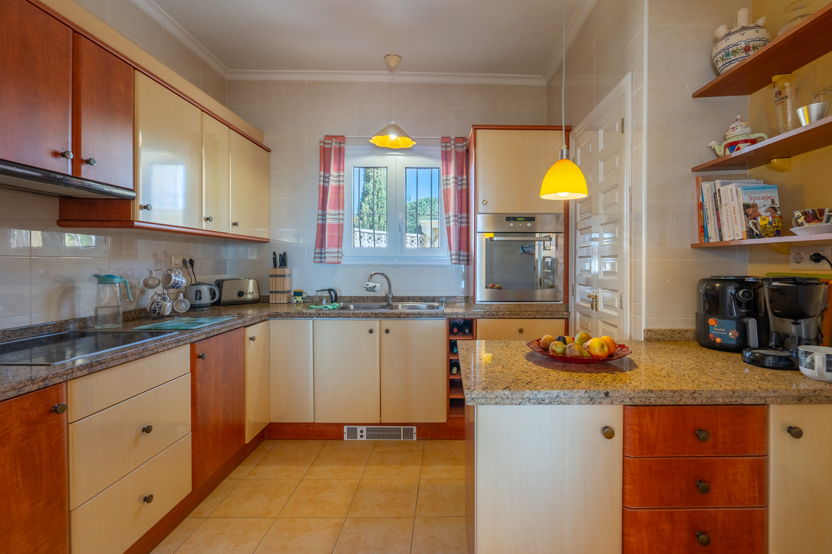 Cuisine équipée avec plans de travail en granit, meubles bicolores en bois et crème, et appareils intégrés. Elle dispose d'étagères ouvertes et d'une fenêtre lumineuse.
