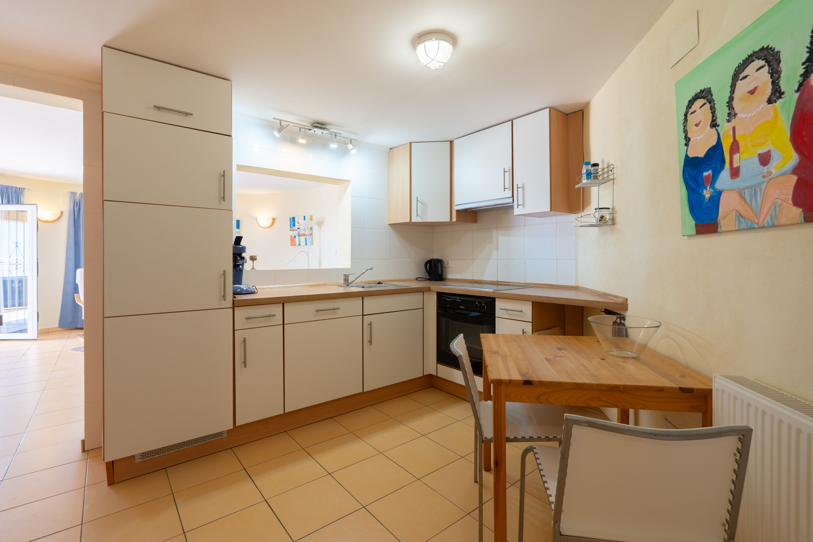 Cuisine fonctionnelle avec placards blancs, plan de travail en bois et coin repas. Elle dispose d'un passe-plat et d'un sol en carrelage aux tons chauds.