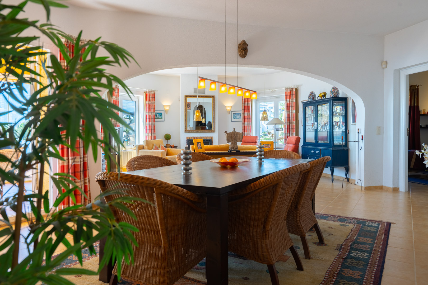 Espace salle à manger lumineux avec table en bois sombre et chaises en osier, relié au salon par une arche. Sols carrelés et lumière naturelle traversante.