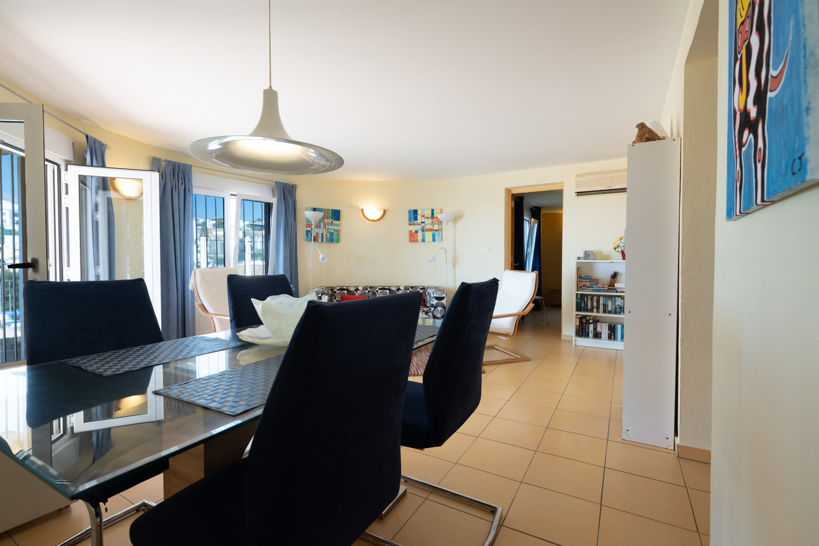 Espace salle à manger lumineux avec table en verre et chaises en velours bleu, intégré dans un salon ouvert avec sol en céramique et climatisation.