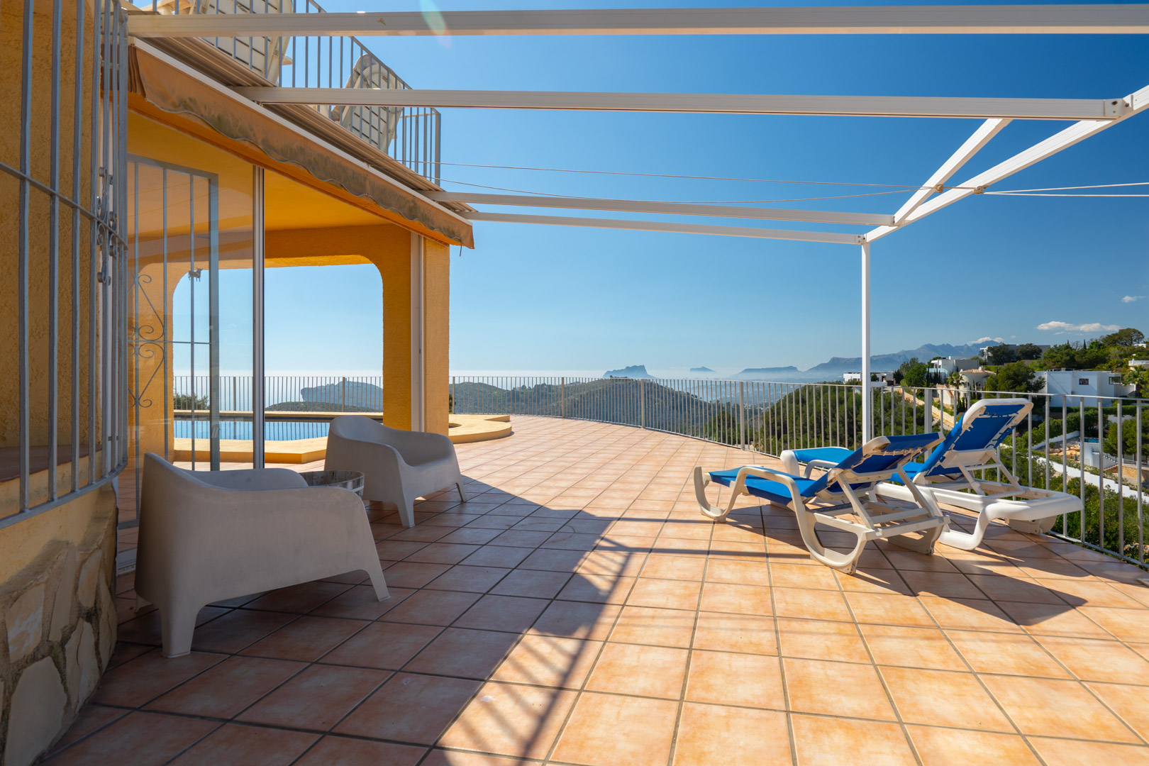 Grande terrasse ensoleillée avec carrelage en terre cuite, pergola métallique et vue panoramique sur la mer et les montagnes environnantes.