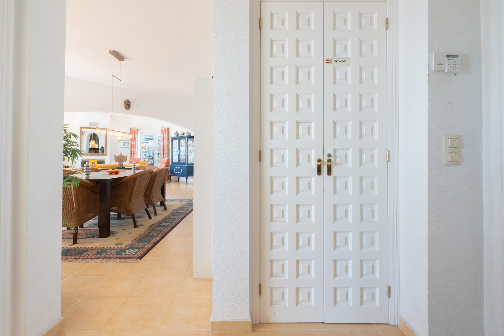 Hall d'entrée avec doubles portes blanches à panneaux ouvrant sur une salle à manger lumineuse avec chaises en osier et sol en terre cuite.
