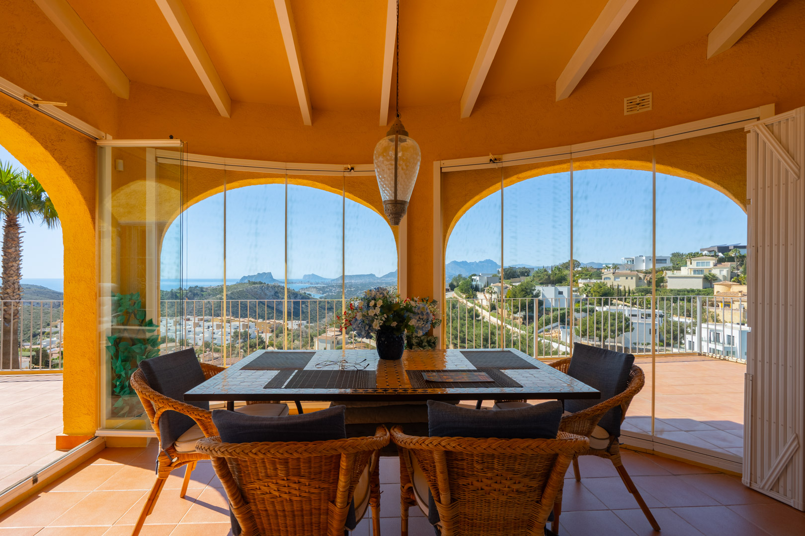 Naya vitrée avec arches traditionnelles et poutres en bois, offrant une vue panoramique sur la mer et les montagnes depuis l'espace repas carrelé.