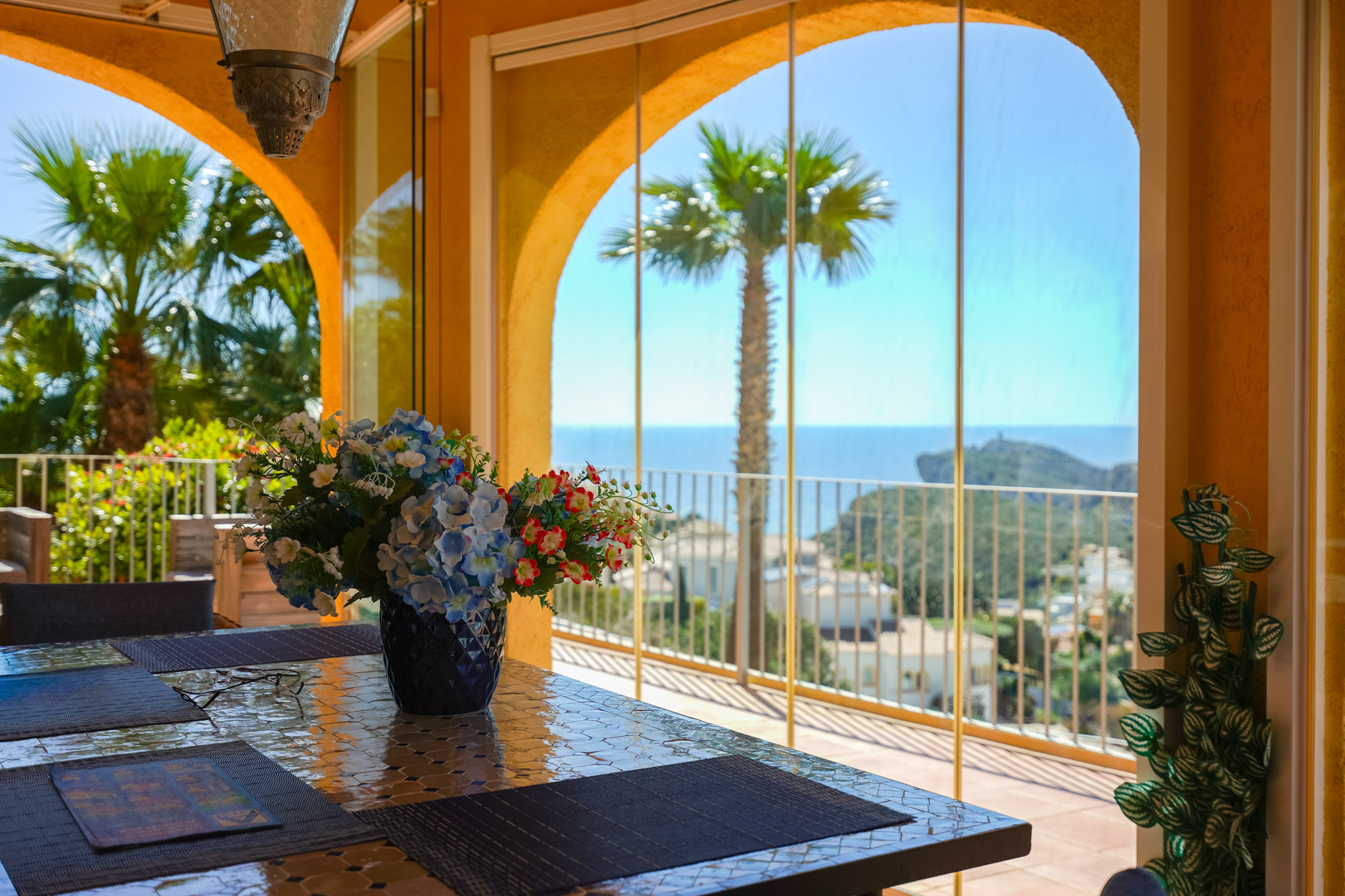 Naya vitrée avec table en mosaïque, offrant une vue panoramique sur la mer et le Cap d’Or à travers des arches traditionnelles et des rideaux de verre.