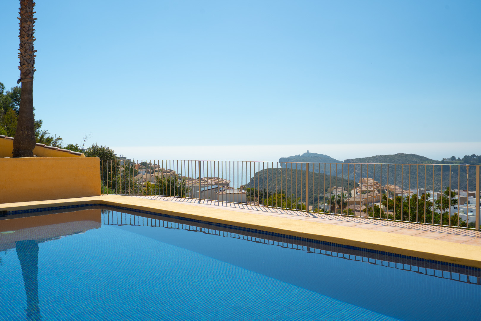 Piscine en mosaïque bleue avec vue panoramique sur la mer et le Cap d’Or à Moraira, bordée d'une terrasse avec garde-corps métallique.