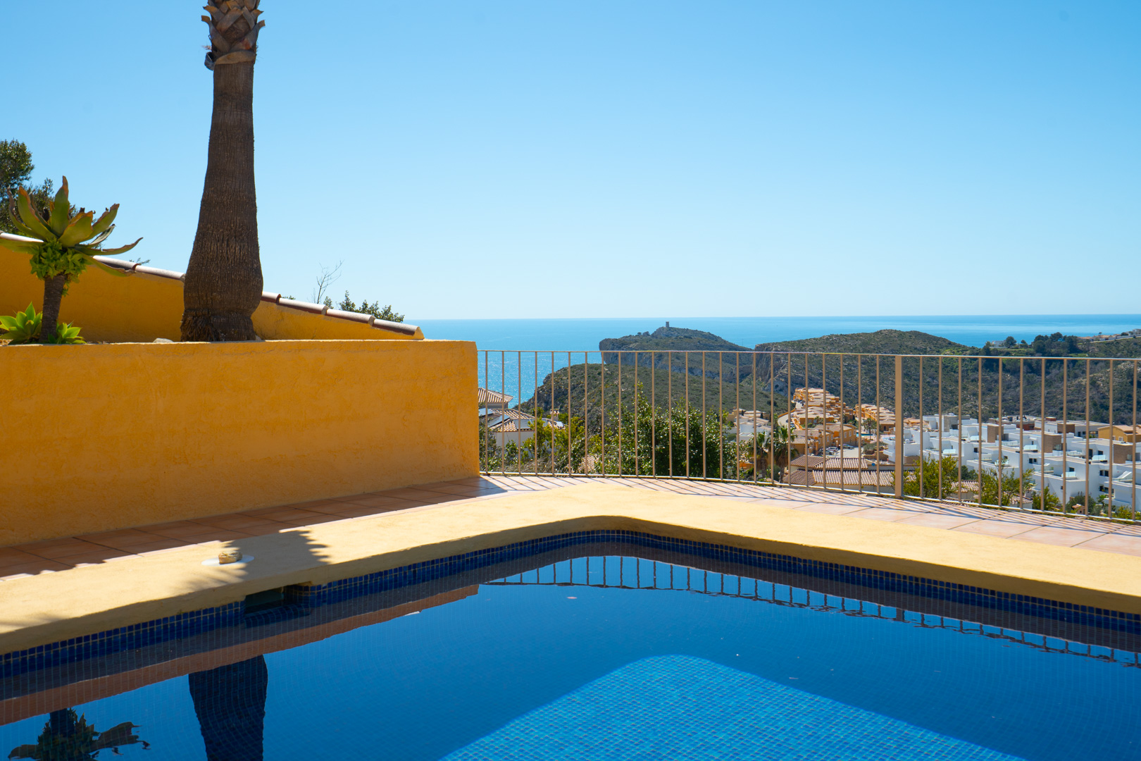 Piscine en mosaïque bleue et terrasse offrant une vue panoramique sur la mer Méditerranée et le Cap d'Or à Moraira, bordée d'une balustrade métallique.
