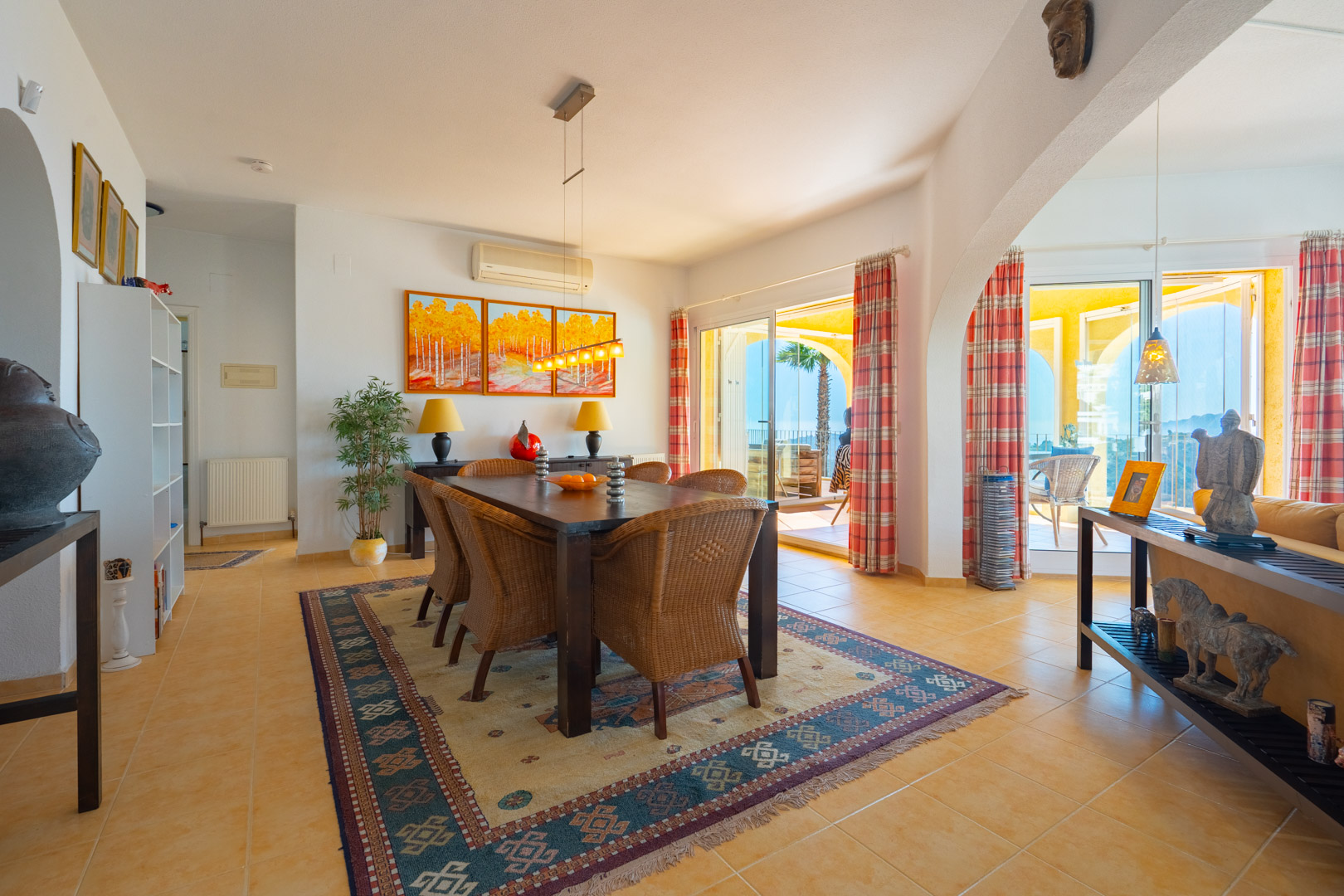 Salle à manger avec table en bois sombre et chaises en osier, ouvrant sur une terrasse couverte arquée avec vue sur mer via de larges baies vitrées.