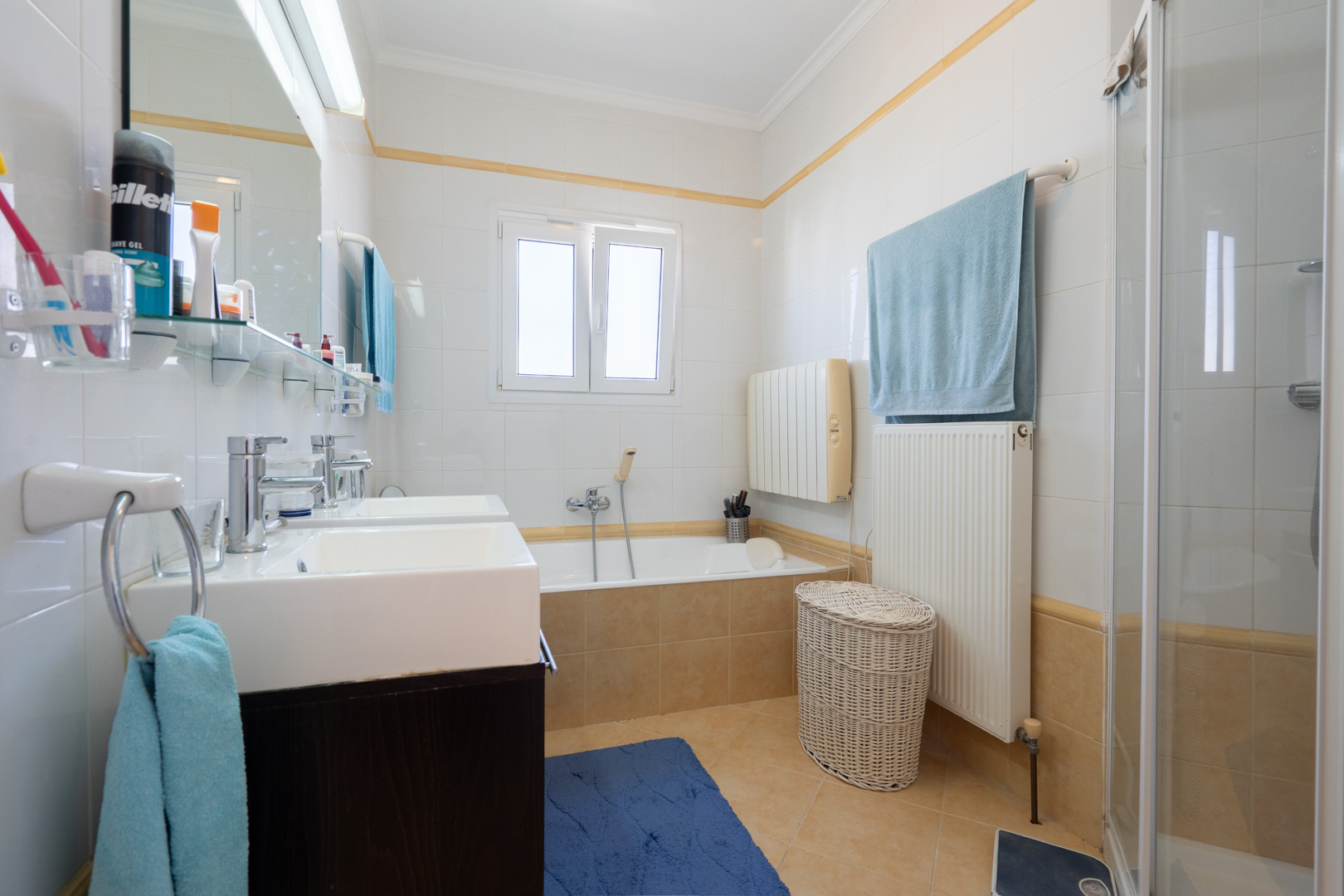 Salle de bain complète avec double vasque, baignoire encastrée, cabine de douche séparée et carrelage blanc avec liseré décoratif.