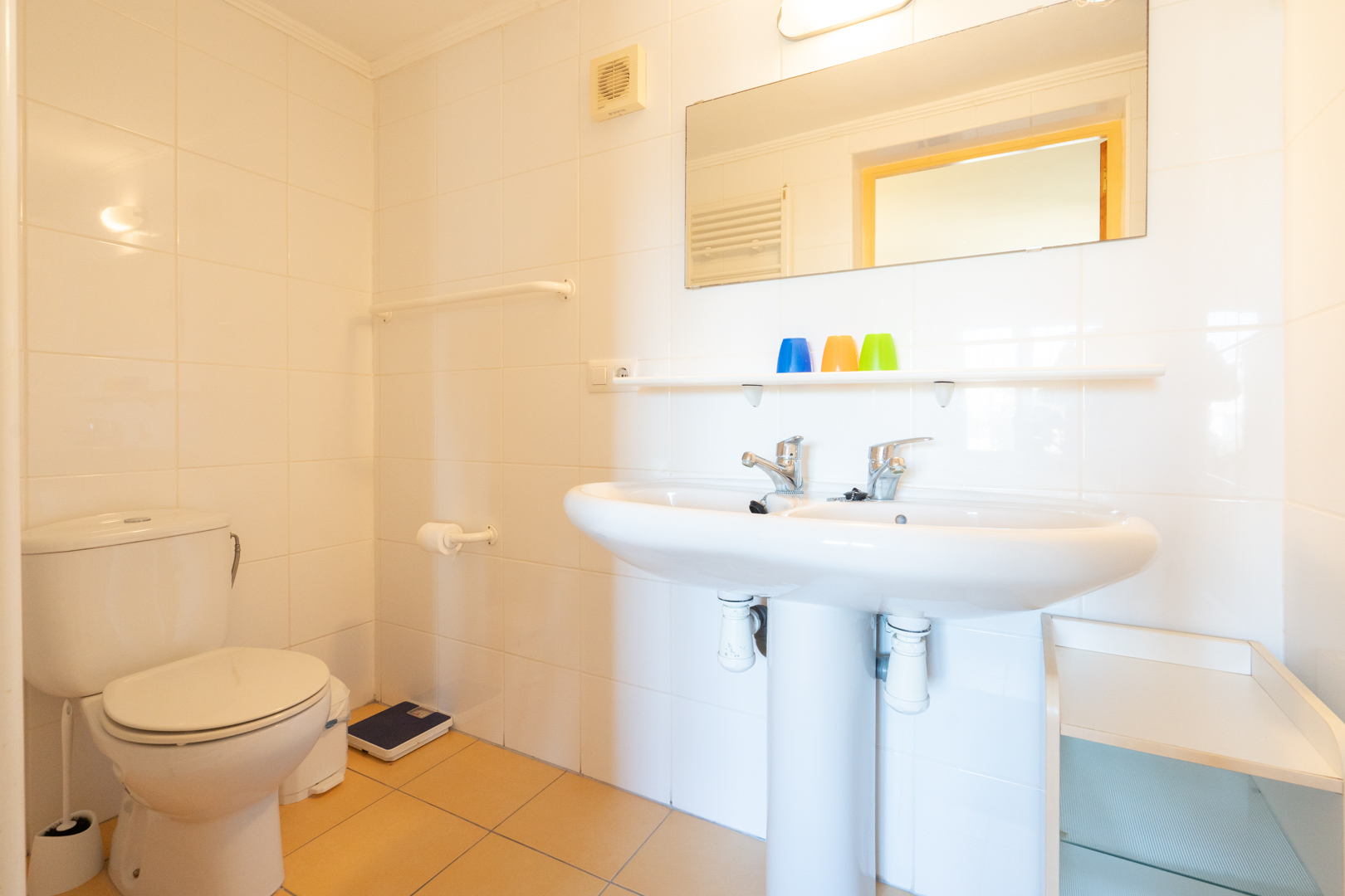 Salle de bain fonctionnelle avec double vasque sur colonne, murs carrelés en blanc et sol en grès. Comprend un grand miroir et des rangements intégrés.