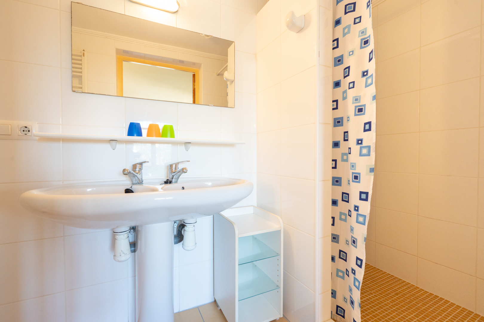 Salle de bain fonctionnelle avec lavabo à double robinetterie, murs carrelés blancs et espace douche avec rideau à motifs et sol en mosaïque ocre.