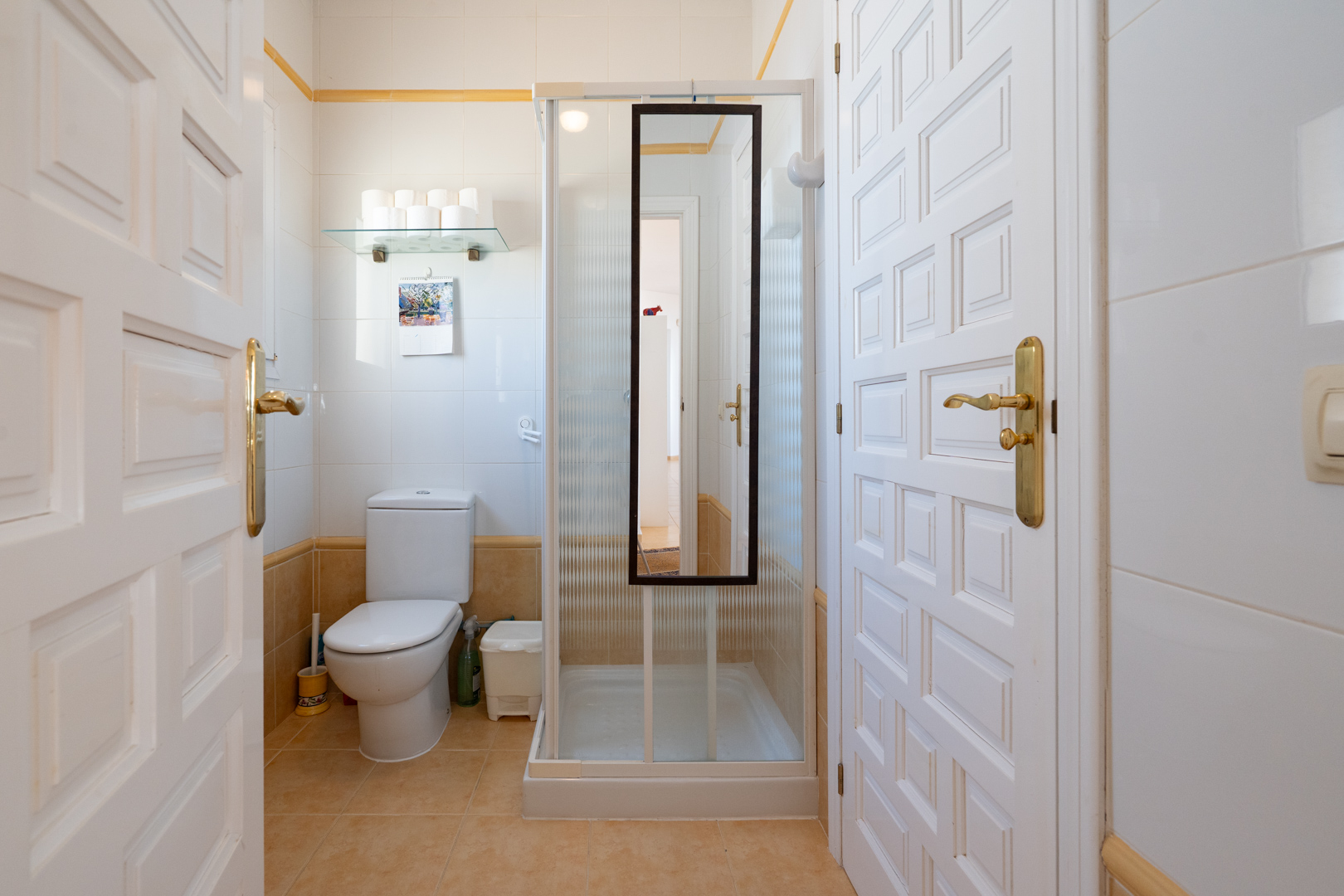 Salle d'eau avec carrelage blanc et ocre, équipée d'un WC et d'une cabine de douche avec miroir, accessible par des portes blanches à panneaux.