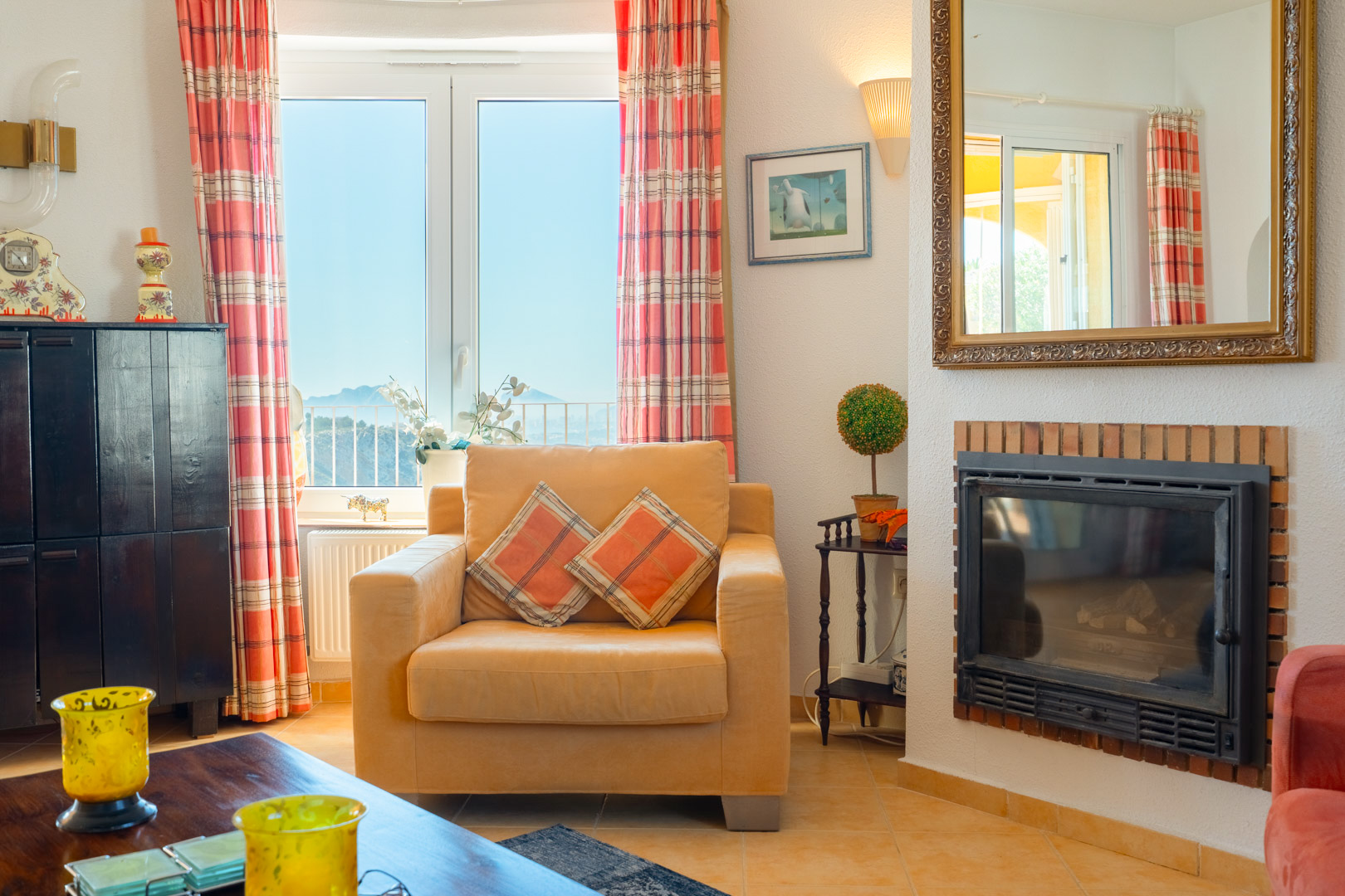 Salon avec cheminée encastrée, fauteuil confortable et fenêtre offrant une vue sur les montagnes. Décoré d'un grand miroir doré et de rideaux à carreaux.