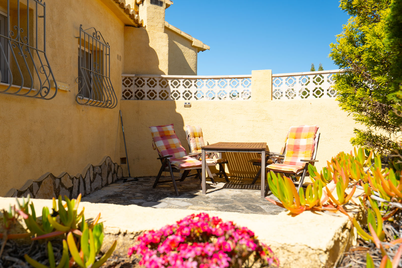 Terrasse extérieure avec sol en pierre naturelle, mobilier en bois et façade ocre ornée de grilles traditionnelles en fer forgé.