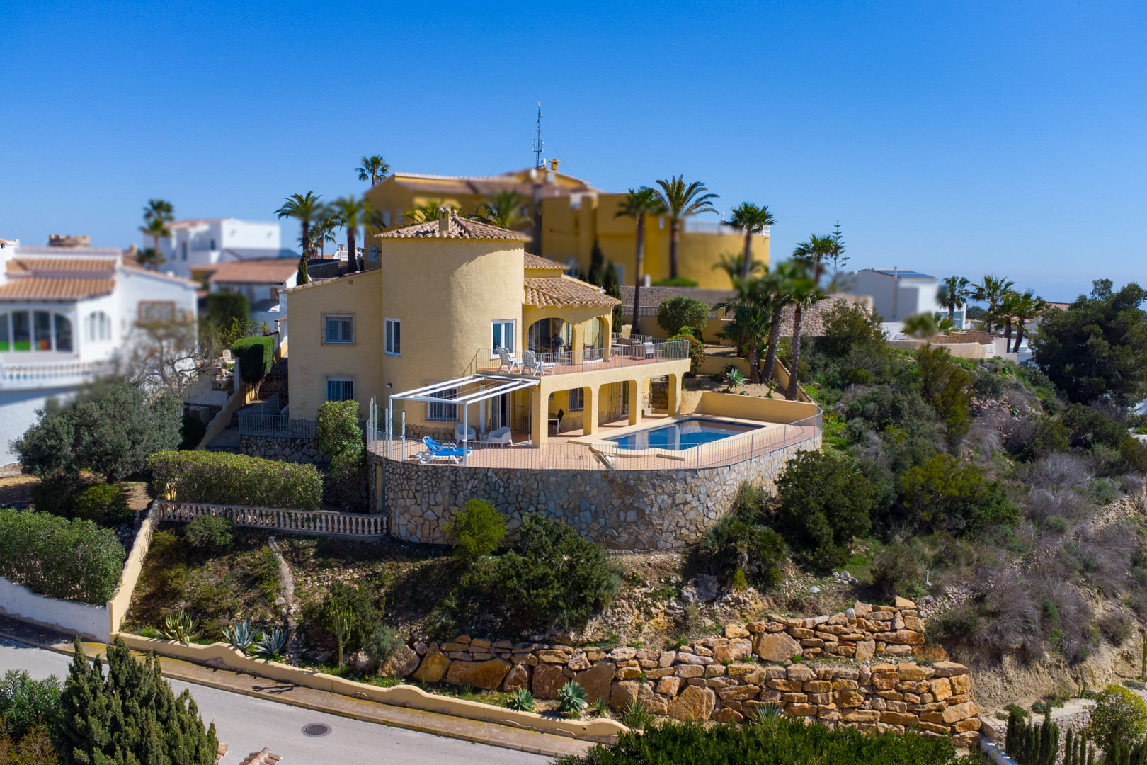 Vue extérieure de la villa avec façade ocre, tour circulaire et piscine rectangulaire située sur une vaste terrasse soutenue par des murs en pierre.