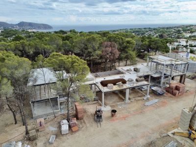 Vue aérienne de la villa en construction, révélant sa structure en béton et les matériaux de chantier. La mer Méditerranée et le paysage boisé sont visibles en arrière-plan.