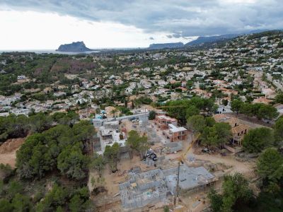 Vue aérienne de villas en construction dans un quartier résidentiel de Moraira, avec grues et fondations visibles, encadrée par la mer et le Peñón de Ifach au loin.